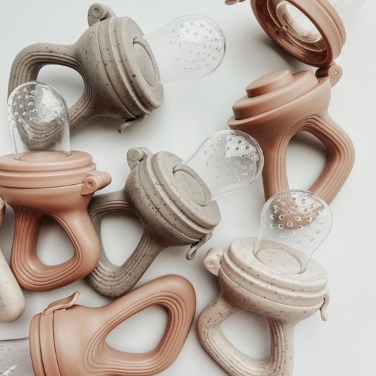 Silicone baby fruit feeders in neutral beige and warm brown tones shown open and closed, highlighting the clear feeding chamber and easy-grip handle.